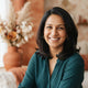 A friendly, professional headshot of a smiling South Asian woman against a warm-toned, blurred background.